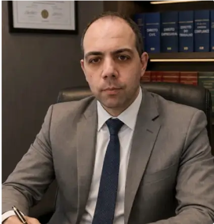 Advogado Trabalhista no Rio de Janeiro Victor Agostini de terno cinza e gravata azul marinho sentado em uma cadeira em um escritório com uma estante de livros atrás